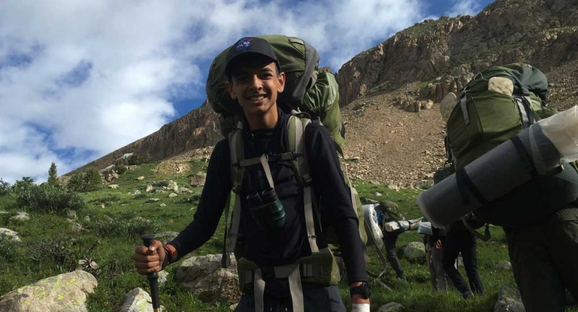 a young person wearing a backpack smiles at the camera amidst a mountainous landscape